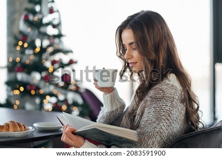 Side view of young cute brunette woman in cozy sweater holding cup coffee or tea and reading interesting book on Christmas tree background. Concept of Christmas atmosphere at home.