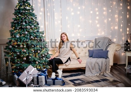 A beautiful lovely woman in a beige sweater sits on the floor against the background of a Christmas tree and decorates a Christmas tree with toys in a spacious room decorated with garlands.