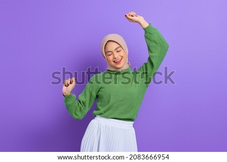 Beautiful young Asian woman in a green sweater raises arms and dances to favorite music, forgot about all problems isolated over purple background Royalty-Free Stock Photo #2083666954