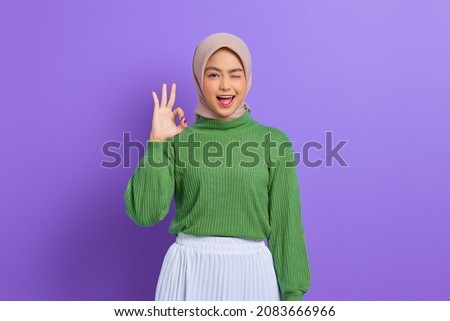 Beautiful smiling Asian woman in green sweater showing okay gesture demonstrates symbol of approval isolated over purple background