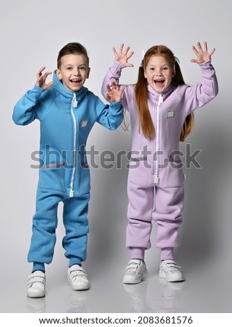 Two laughing, frolic kids boy and girl in blue and pink jumpsuits stand holding hands up, showing beast claws, playing, scare us over light background