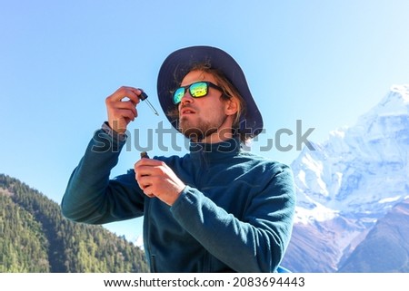 Young caucasian male with a beard wearing a fleece, a hat and sunglasses taking CBD oil from a dripper high up in the Himalaya mountains. Sports enhancement supplement for trekking in high altitude.