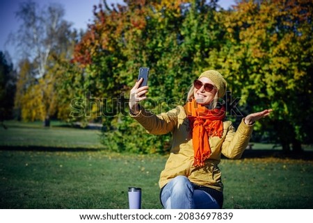 Cheerful girl in bright clothes and sunglasses sitting on park bench and taking selfie on smartphone against autumn trees. Attractive woman makes hand gestures on the background of nature. Copy space.