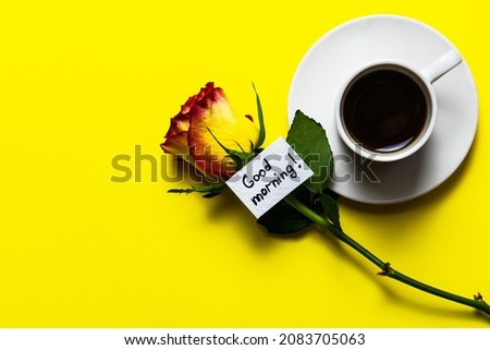 a cup with a rose and good morning wishes on a yellow background. Good morning is written on a white sheet, which is on a yellow background next to a rose and a white cup of coffee. Selective focus.