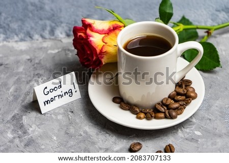 a cup with a rose and good morning wishes on a gray background. Good morning is written on a white sheet, which is on a gray background next to a rose and a white cup of coffee. Selective focus.