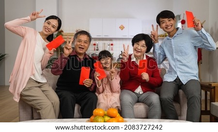cheerful asian extended family looking at camera and making thumb victory hand signs while taking burst shots with lucky money at home on chinese new year