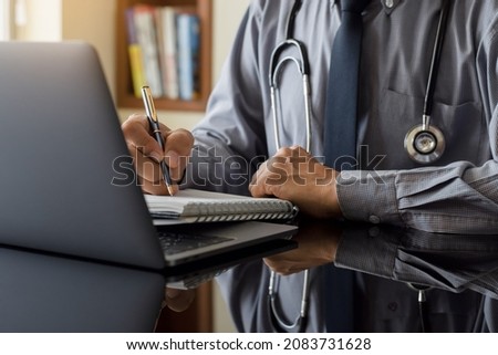 Male doctor writing information on notebook work on laptop computer at medical clinic.  Royalty-Free Stock Photo #2083731628