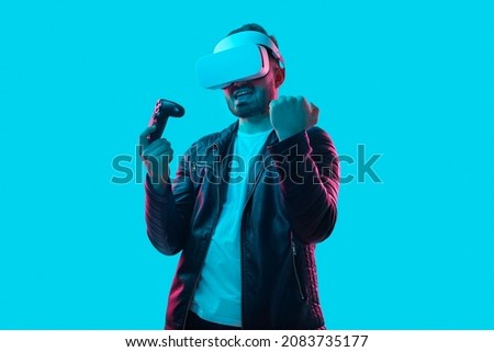 Excited male gamer with controller and in VR goggles celebrating win, while playing videogame in cyberspace on blue background in studio with pink neon light