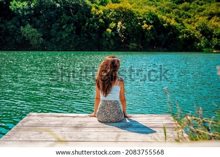 Rear view A young beautiful woman is sitting on a wooden pier on a delightful azure mountain lake surrounded by forest