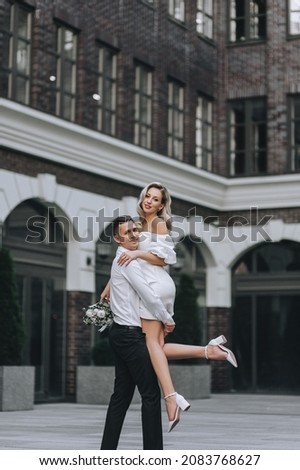 A stylish, cheerful groom holds in his arms a beautiful, young, curly blonde bride in a white dress, dancing against the backdrop of the city and buildings. Wedding photography of the newlyweds.