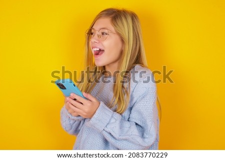 caucasian kid girl wearing blue knitted sweater over yellow background holding a smartphone and looking sideways at blank copyspace.