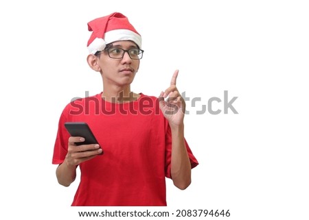 Portrait of excited Asian man in red t-shirt with santa hat standing against white background, pointing away with his finger while using mobile phone. Isolated image