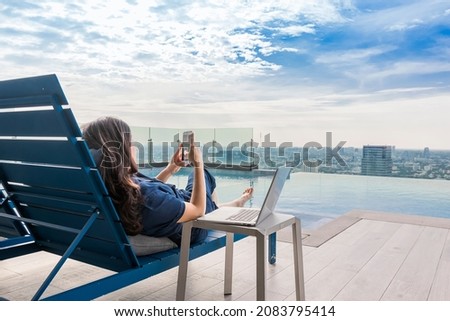 Summer day lifestyle asian woman relax and chill near luxury swimming pool. Asian woman using app on a smartphone and working on a laptop and Blurred background skyscraper, Beautiful sky and clouds. Royalty-Free Stock Photo #2083795414