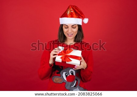 Young woman in a christmas sweater and santa hat on a red background with a gift box in anticipation of the new year
