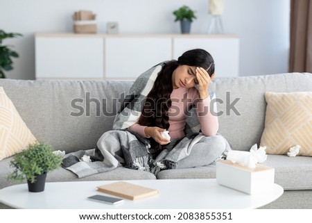 Despaired young indian lady in blanket, suffer from flu and headache, sits on sofa in living room interior with napkins and medication. Home treatment, feeling unwell, cold and covid-19 quarantine Royalty-Free Stock Photo #2083855351