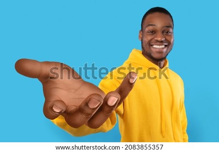 Happy African American guy showing big outstretched hand, offering help, taking or giving something, reaching out for support on blue studio background. Selective focus Royalty-Free Stock Photo #2083855357