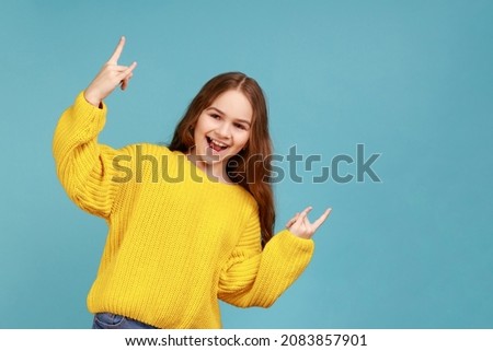 Portrait of happy crazy little girl doing rock and roll gesture, showing cool rock sign with hands, wearing yellow casual style sweater. Indoor studio shot isolated on blue background.