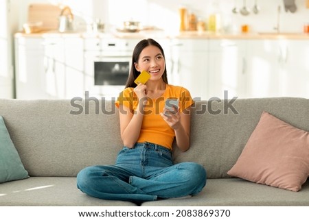 Online shopping concept. Dreamy asian woman with smartphone and credit card sitting on couch, thinking about internet purchases or food delivery, free space
