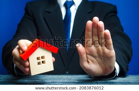 The man holds the house and makes a stop gesture. Bank Refusal to provide a mortgage. Refusal to provide legally guaranteed housing. Confiscation pledged property. Denial of insurance payment. Royalty-Free Stock Photo #2083876012