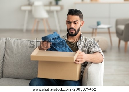 Bad delivery service concept. Angry dissatisfied Arab man unpacking cardboard box, receiving wrong item, feeling upset with parcel at home. Shipping mistake, terrible online order Royalty-Free Stock Photo #2083879462