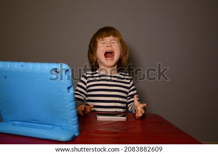 Little boy three years old. Sitting at the table by the blue tablet - the child's emotions on content. The kid laughs. Tablet in a child's protective case.