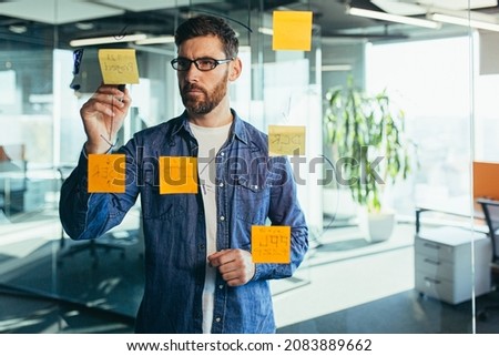 focused and thinking businessman writing on colorful notes attached to a glass wall, the entrepreneur creates a visual to-do list, business goals for future careers, daily goals