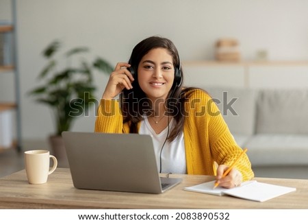 Distance education. Happy arab woman in headphones sitting at desk, using laptop and writing in notebook, smiling at camera. Female student taking notes, studying online at home