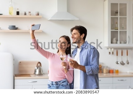 Smiling millennial european couple with glasses of wine on date at home taking photo on smartphone on kitchen interior. Photo at anniversary, video call and holiday, online livestream for blog, indoor