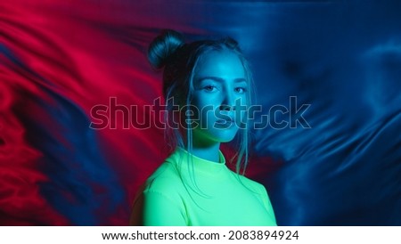 Modern young teen girl blowing gum bubble in changing colorful light. Confident female model in iridescent multicolors of lighting on background with triangular neon lamp.