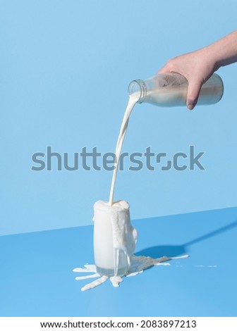 Woman hand pouring milk from a bottle into the glass, minimalist on a blue background. Over-spilling milk on the blue-colored table. Royalty-Free Stock Photo #2083897213