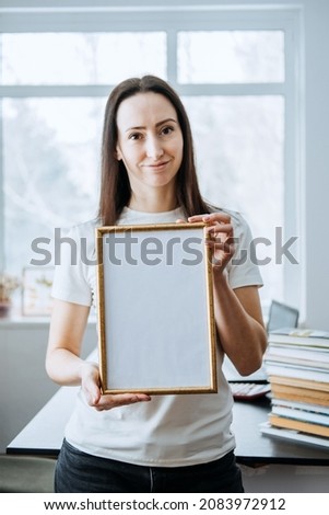 Frame picture, poster, diploma, certificate mockup in female hands. Young brunette woman holding empty blank wooden photo frame on table with laptop and books home office background