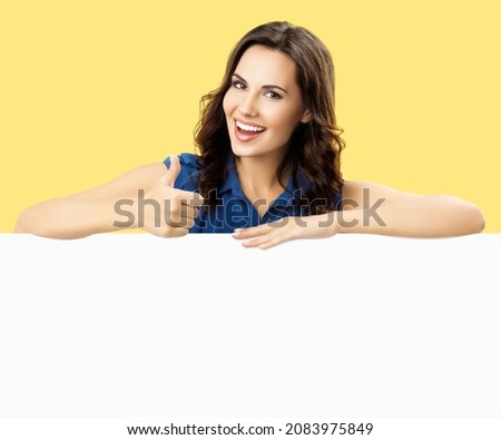 Happy smiling brunette woman in blue dress cloth, showing thumb up hand gesture, standing behind, peeping from blank banner mock up ad billboard signboard with copy space over yellow color background.