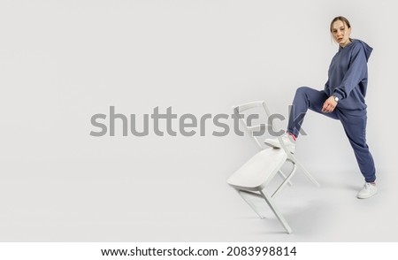 Full length portrait of young sporty fitness women in blue cotton hooded sweatsuit casual outfit playing with white chair. Studio shot of blonde Hoody woman with long hair in hoodie activewear set. Royalty-Free Stock Photo #2083998814