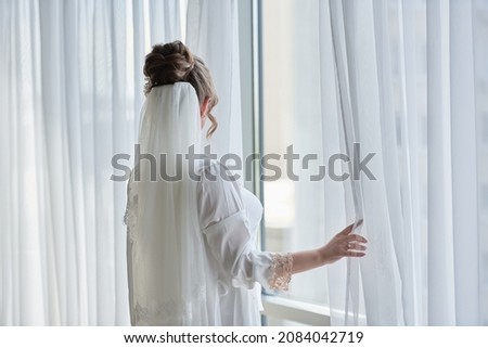 Portrait of beautiful bride in white silk dressing gown with curly hairstyle and veil standing near window in bedroom, copy space. Bride's morning preparation. Back view