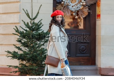 Christmas street style portrait of young beautiful woman in red beret walking in European city for winter holidays. Stylish model with curly hair is doing New Year's shopping. Cozy.