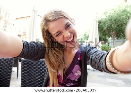 Close up portrait of a young attractive woman holding a smartphone digital camera with her hands and taking a selfie self portrait of herself winking at he camera. Travel and technology outdoors.