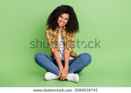 Full size photo of pretty volume hairstyle millennial lady sit wear spectacles yellow outfit jeans sneakers isolated on green background