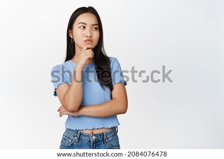 Troubled asian girl thinking, frowning and looking pensive aside, having problem, standing over white background