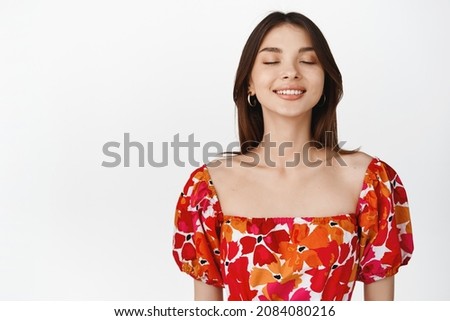 Portrait of beautiful brunette woman close eyes and dreaming, smiling while imaging something, standing in floral dress over white background