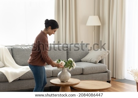 Smiling young indian ethnicity woman homeowner putting beautiful flowers in vase on coffee table in modern living room, enjoying decorating own apartment, improving styling interior on weekend. Royalty-Free Stock Photo #2084086447