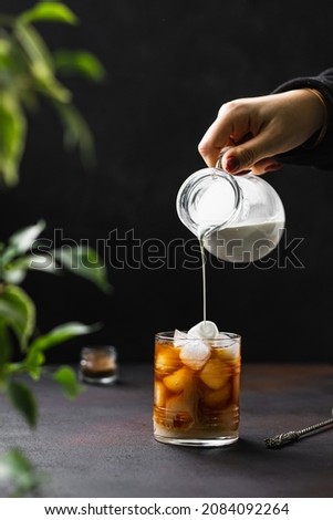 Iced coffee in glasses with milk pouring from pitcher by hand, green plant branches at background, copy space, square crop. Summer refreshing beverage ice coffee drink concept. Dark photo