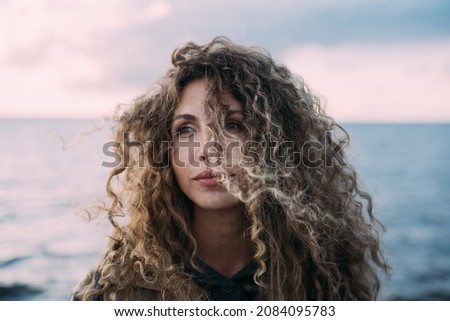 Portrait of young happy smiling woman on the coast in the summer. A woman with curly hair resting day on the beach in windy weather. Close up picture.