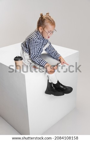 Model wearing glasses and checkered shirt. Cute little girl with laptop and coffee cup pointing down isolated over gray background. Concept of childhood, education, motherhood. Copy space for ad