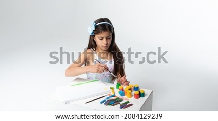 beautiful girl with a light blue bow on her head, holding her brush, painting a canvas with green paint, next to colorful paints.