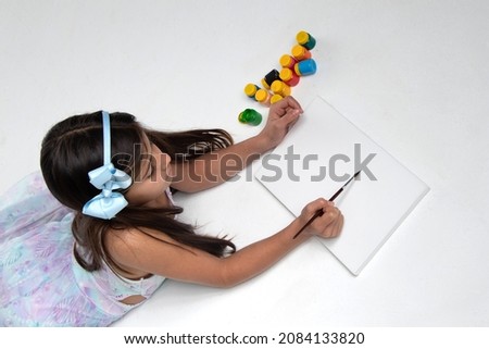 beautiful girl lying with a light blue bow on her head, holding brush, painting a blank canvas, next to colorful paints, photo view from above.