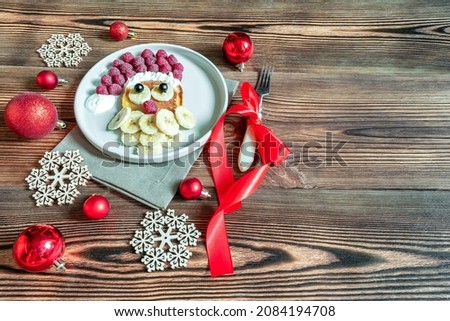 Christmas Santa Claus face shaped pancake with sweet fresh raspberry berry and banana on plate on wooden background for kids children breakfast. xmas food dessert with new year decorations, red balls