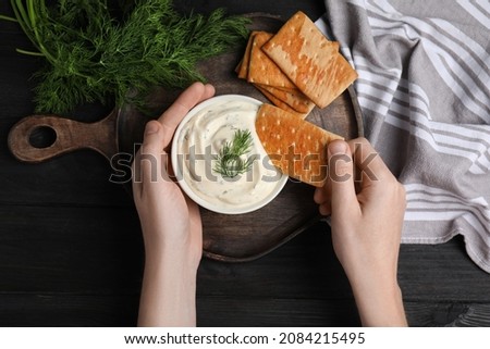 Woman dipping cracker into tasty creamy dill sauce at black wooden table, top view