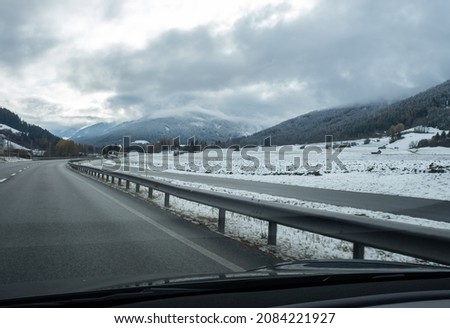 A shot on the move from behind the windshield of an electric car with snow-covered alps mountains. Cold cloudy autumn day. POV first person view shot on a asphalted mountain road. Selective focus.