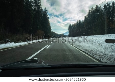 A shot on the move from behind the windshield of an electric car with snow-covered alps mountains. Cold cloudy autumn day. POV first person view shot on a asphalted mountain road. Selective focus.