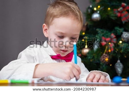 Cute serious boy, in a white shirt and pink bow tie, writing letter to Santa Claus on Christmas eve at home.